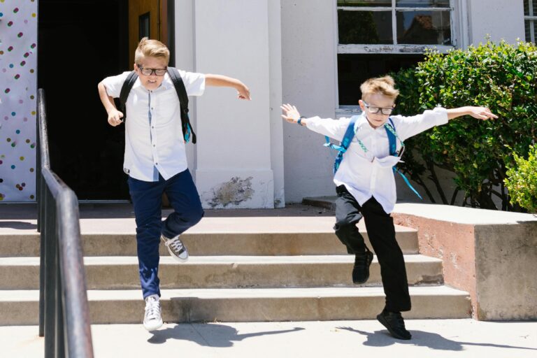 Two boys with backpacks energetically jumping down concrete steps outside a school.